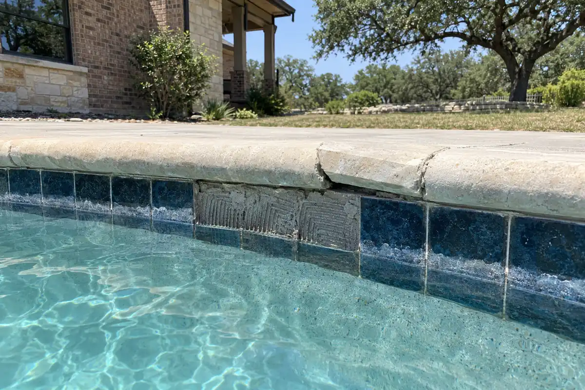 Popped waterline tiles exposing the mortar bed under a cracked, lifted limestone coping stone on a full pool